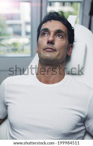 Handsome male patient dressed in the white t-shirt seated on the medical chair in the cabinet of aesthetic clinic