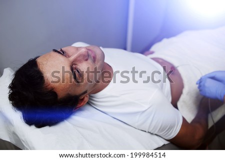 Handsome male patient dressed in the white t-shirt lying on the medical chair at body mesotherapy in the cabinet of aesthetic clinic