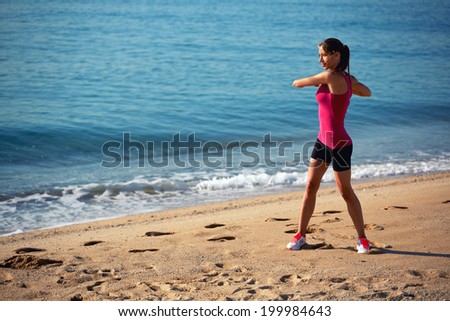 Sports girl dressed in pink t-shirt and black shorts doing workout standing on the sand on beautiful sea background, morning jog