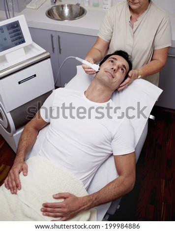 Handsome male patient dressed in the white t-shirt lying on the medical chair at facial mesotherapy looking to the female doctor in the cabinet of aesthetic clinic