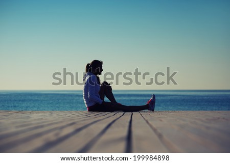 Athletic female runner resting after the stretching exercise seated on the beach and looking away, female runner seated on the wooden pier looking away
