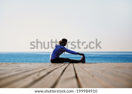 Beautiful athletic woman sitting on the wooden pier doing stretching exercise before the morning jog on the sea background