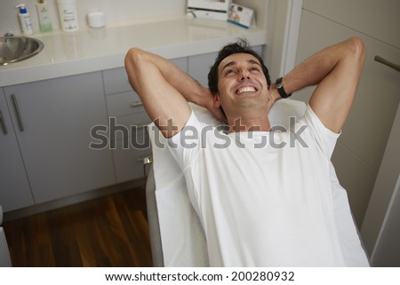 Happy satisfied patient lying in the medical chair of aesthetic clinic, attractive man in white t-shirt relaxing in the cabinet of beauty salon