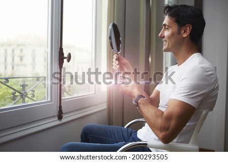 Attractive man looking at himself in a hand mirror after rejuvenating in aesthetic clinic, male patient seated across the window
