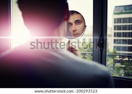 Handsome man looking at himself in a hand mirror after the plastic surgery in aesthetic clinic, patient after skin-juventation procedure