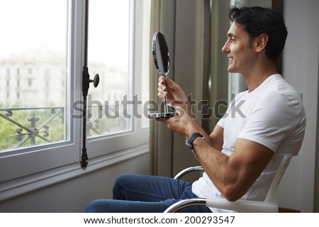 Handsome man  in the white t-shirt sits across the window and looking at himself in a hand mirror after plastic surgery