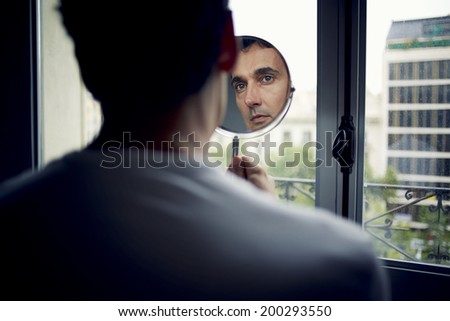 Handsome adult man looking at himself in a hand mirror after the plastic surgery in aesthetic clinic