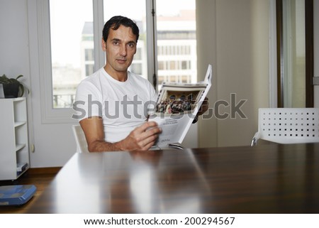 Beautiful man in a white shirt reads journal sitting at a wooden table in the big clean room