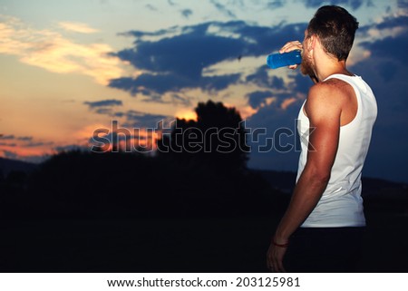 Attractive runner with muscular body enjoying amazing sunset drinking water, runner resting after intensive evening jog, health lifestyle and fitness concept