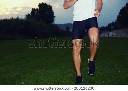 Athletic muscular runner limber up lifting his feet up before the evening jog, workout outside, sportsman doing exercise in the park at evening training jog