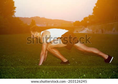 Athletic muscular runner in white t-shirt ready to start standing on the grass on the beautiful sunset background, male runner at evening jog in the park, health lifestyle and fitness concept