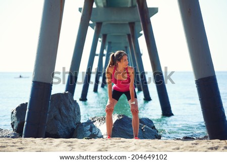 Attractive woman with beautiful muscular body resting after intensive run on the beach, female runner resting on the beautiful sea background, fitness and healthy lifestyle concept