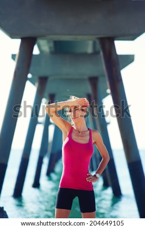 Female runner with beautiful muscular body wipe the forehead with the hand, athletic attractive woman resting on the beach after morning run, fitness and healthy lifestyle concept