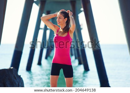 Beautiful girl in bright sportswear resting on the beach after intensive morning run, female runner taking break on the beautiful sea background, fitness and healthy lifestyle concept
