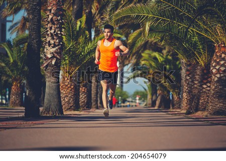 Muscular athletic man running on the jogging track, athletic male jogger running along beach road with palm trees, evening run at beautiful sunset, fitness and healthy lifestyle concept