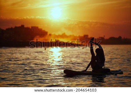 Silhouette of standup paddle boarding performed by beautiful athletic girl, orange sunset reflected in the sea, yoga at perfect sunset, spiritual meditation and relax
