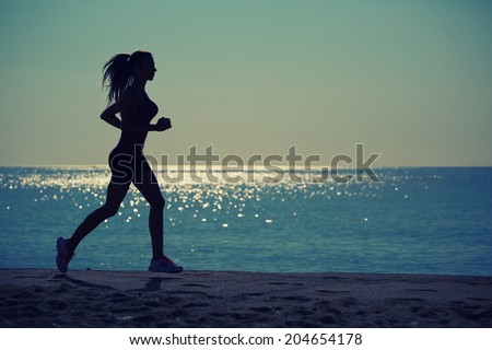 Silhouette of athletic female jogger with beautiful muscular figure running on the beach, female runner run along the sea, morning run on the beach, fitness and healthy lifestyle concept