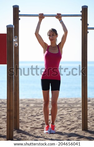 Beautiful athletic woman with muscular body standing on the beach at horizontal bar on amazing sea background, workout outdoor, fitness and healthy lifestyle concept