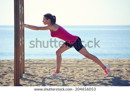 Attractive sporty woman with muscular body doing stretching exercise on the horizontal bar, workout outdoor, female runner on the beach, fitness and healthy lifestyle concept