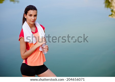 Female attractive runner with the bootle of water in the hands standing on beautiful lake background, jogger resting after run,fitness and healthy lifestyle concept