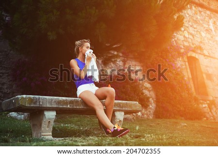 Beautiful sporty girl seated in the bench clean his face with towel, tired blond runner resting after evening jog, fitness and healthy lifestyle  concept