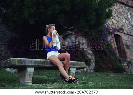 Beautiful female runner seated in the bench clean his face with towel, tired blond runner resting after evening jog, fitness and healthy lifestyle  concept