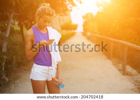Blond sporty girl looking to the telephone screen taking break after evening run, female runner with towel in shoulder holding bottle of water in the hands, fitness and healthy lifestyle  concept