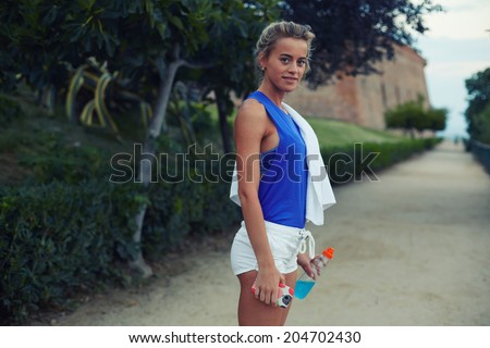 Female runner with beautiful figure standing in big green park at evening jog, sporty girl with bottle of water in the hands, fitness and healthy lifestyle concept