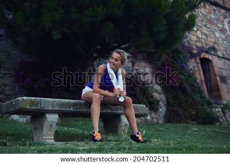 Attractive female runner resting in the park seating on the bench, evening run in the park, female runner with towel and bottle of water, fitness and healthy lifestyle  concept