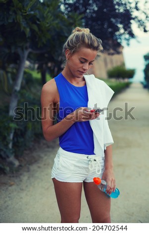 Attractive female blond runner standing with telephone in the park, evening jog and sport outside, fitness and healthy lifestyle concept