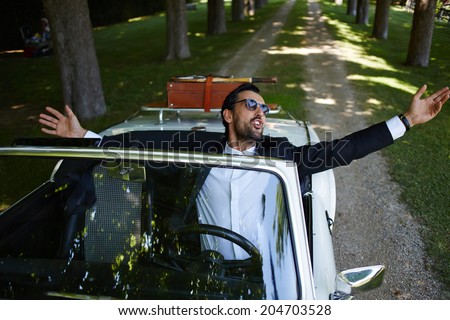 Successful stylish man with arms raised and feeling of freedom sitting behind the wheel of cabriolet classic luxury car, lifestyle and successful business concept
