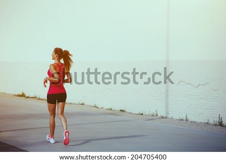 Attractive sporty girl with muscular body on the morning jogging at sunny day, female athletic jogger runs along pastel color wall, healthy lifestyle and fitness concept