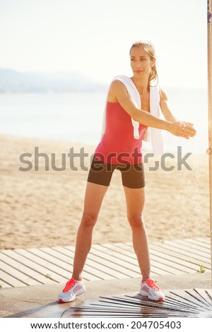 Athletic woman with beautiful figure standing near a beach shower and washes hands, female jogger resting after intensive morning jog on the beach, healthy lifestyle and fitness concept