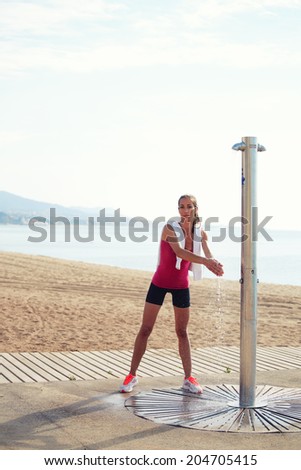 Attractive female runner standing near a beach shower and washes hands, female jogger resting after intensive morning jog on the beach, healthy lifestyle and fitness concept