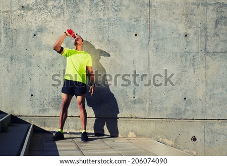 Young muscular build man with running armband on the arm drinking water of bottle, attractive athlete resting after workout outdoors, fitness and healthy lifestyle concept