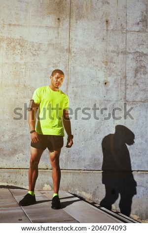 Healthy African American runner with running armband on the arm resting tired after evening training, beautiful male runner resting standing on concrete wall background, healthy lifestyle concept