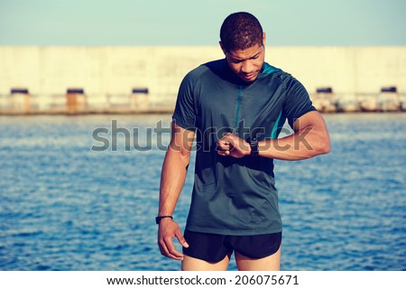 Running athlete man looking at smart watch having rest after jogging outdoors, healthy african american runner resting tired after evening training standing in the sea port at sunset, fitness concept