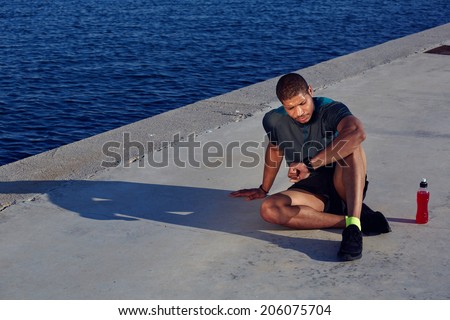 Attractive male runner looking at smart watch having rest after jogging outdoors, healthy african american man resting tired after evening training seated in the sea port at sunset, fitness concept