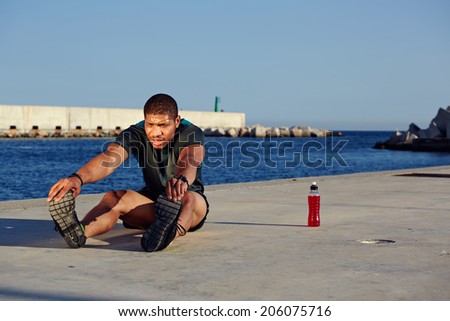 Attractive muscular build runner stretching his legs seated on sea port background, exercising on a sunny day and workout outdoors, fitness and healthy lifestyle concept