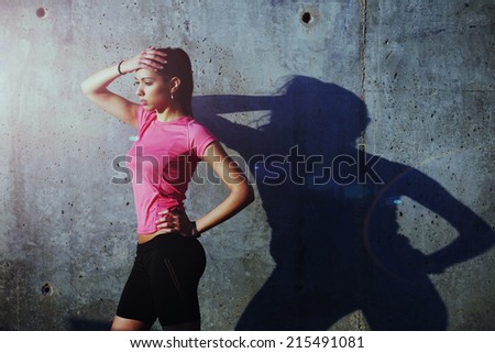 Beautiful athletic girl resting after intensive jogging outdoors, tired after evening run attractive female runner resting on concrete wall background at sunny evening, healthy lifestyle concept