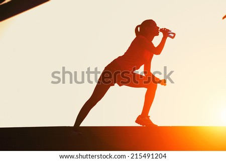 Silhouette of attractive female runner drinking water on beautiful sunset background, jogger with muscular body taking break after intensive evening training, sports advertising and healthy lifestyle