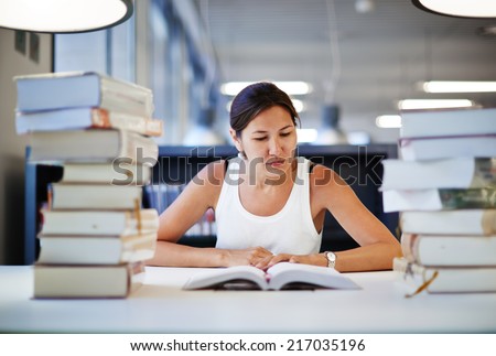 Young college student girl sitting at the library desk and reading the book, preparing to the exams in high school young asian student seated with pile of books in bright library