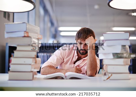 Young male college student sitting at the library desk with two big pile of books, bored and tired to study high school student boy reading the book, preparing to the exams in library