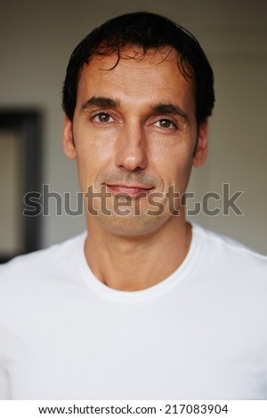 Portrait of handsome man with tanned skin with positive facial emotion looking to the camera indoors, adult attractive man in white shirt putting the hands on the side smiling to the camera