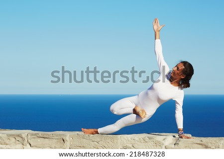 Brunette woman with slim body stand in yoga pose on blue sea background, healthy young woman meditating yoga enjoying sunny evening at summer, practicing yoga at sunny day, feeling healthy