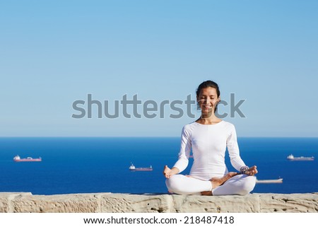 Attractive brunette girl smiling seated in yoga pose on beautiful sea background, healthy woman smiling during yoga training outdoors, happy woman practicing yoga enjoying sunny day at summer