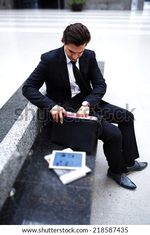 Rich serious businessman on the way to conference looking for something in his work bag, successful businessman in black suit seated on the train station bench with newspaper and tablet