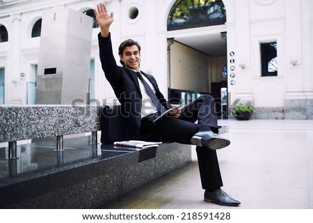 Handsome man in black suit seated on the bench of big railway station say hello to someone raising the hand, confident business man on the way to work waiting for the train in big train station