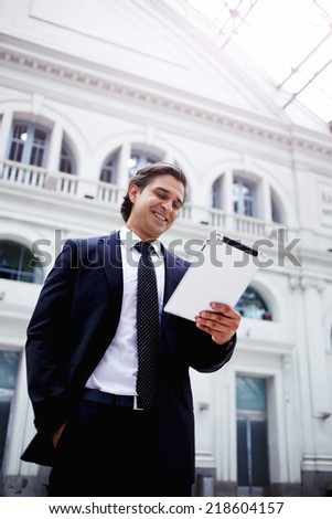 Happy rich businessman using his PC tablet standing in big light hall of railway station, smiling attractive business man using his digital tablet while waiting next train in the big railway station