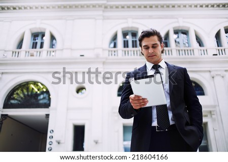 Mid adult businessman standing in big light hall holding tablet pc in the hands, happy business man reading news standing in the big railway station on the way to work, business people and finance
